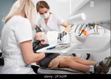 Kleiner Junge, der im Zahnstuhl sitzt, während zwei weibliche Zahnärzte Zähneputzen überprüfen. Zahnarzt untersucht junge Zähne mit zahnmedizinischen Instrument. Konzept der Kinderzahnheilkunde und Zahnpflege. Stockfoto