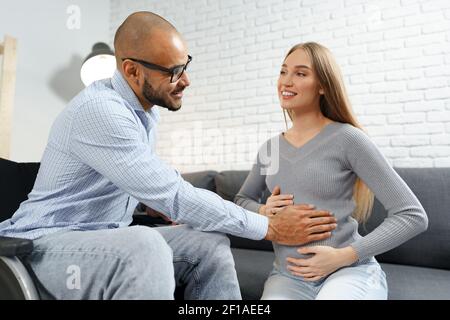 Behinderter afroamerikanischer Mann, der den Bauch seiner Schwangeren berührt kaukasische Frau, während sie auf dem Sofa zu Hause entspannen Stockfoto