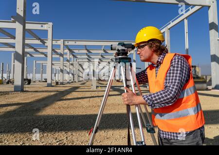 Bauarbeiter in Yellow HardHat vor dem Industriegebäude im Bau. Bauarbeiter mit Tachometer vor Ort im Bau Stockfoto