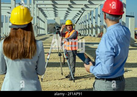 Erwachsener Landvermesser mit Tachometer bei der Kamera. Bauarbeiter in Gelb HardHat . Baugewerbe-Team auf der Baustelle. Stockfoto