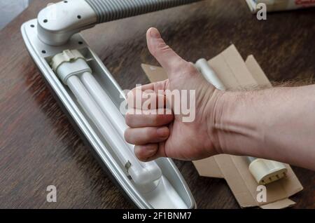 Die Hand eines Mannes zeigt eine Geste mit Daumen nach oben. Eine graue Tischleuchte mit einer neuen kompakten Leuchtstofflampe und einer alten Lampe auf der Rückseite eines Holztisches. Ersetzen Stockfoto