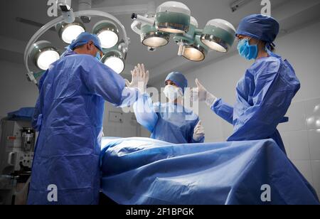 Ärzte stehen nach erfolgreicher operativer Operation beim Patienten. Plastischer Chirurg, der dem Assistenten hohe fünf gibt. Zweiter Assistent macht Daumen hoch Geste. Konzept der Medizin und erfolgreiche Teamarbeit. Stockfoto