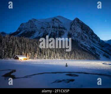 Winternachtszene auf gefrorenem See, der von Schnee bedeckt ist, teilweise von Bootshäusern beleuchtet, aufgenommen in Lake Louise, Alberta, Kanada Stockfoto