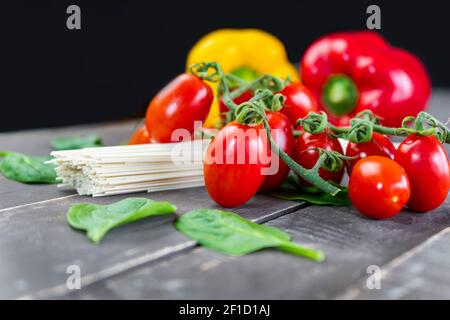 Nahaufnahme von frischem Bio-Gemüse mit roher Pasta auf Holztisch.Kirschtomaten, Spinatblätter und bunte Paprika auf dunklem Hintergrund. Stockfoto