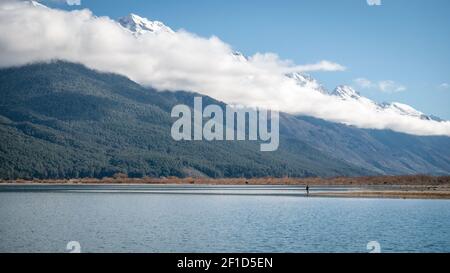 Mann, der am Ufer des Sees steht, mit Blick auf große Berge, die von einer niedrigen Wolke umhüllt sind. Gedreht in Glenorchy, Neuseeland Stockfoto