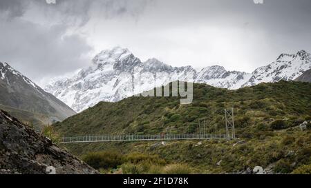 Blick auf den Berg mit Mann, der im Vordergrund die Swing Bridge überquert, aufgenommen im Aoraki/Mt Cook National Park, Neuseeland Stockfoto