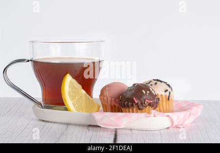 Tasse Tee mit Zitronenkeil, begleitet von kleinen Cupcakes mit verschiedenen Geschmacksrichtungen und Farben. Tablett mit rosa und weiß karierten Servietten platziert. Stockfoto