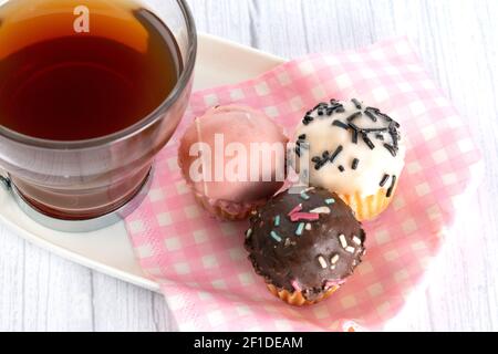 Tasse Tee, begleitet von kleinen Cupcakes mit verschiedenen Geschmacksrichtungen und Farben. Platziertes Tablett mit rosa und weiß karierten Servietten.. Stockfoto