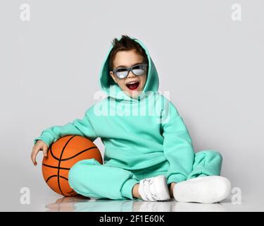 Happy Screaming frolic Kind Junge in modernen grünen, mint Farbe Sportswear Hoodie, Hosen und Sneakers sitzen mit Basketball