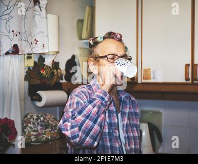 Ältere schwedische Frau in Haarwickler trinken aus Becher in der Küche. Konzept des Lebensstils Stockfoto