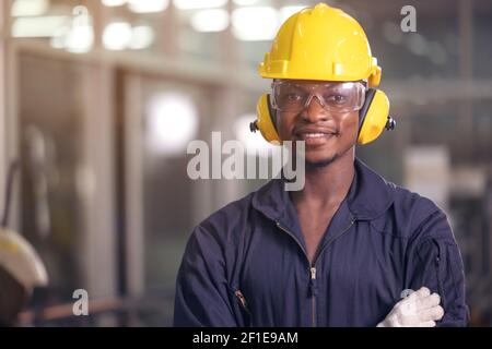 Portrait von fröhlichen schwarzen Arbeiter trägt schützende Kopfhörer posiert suchen Bei der Kamera und genießen Sie die Arbeit im Hintergrund Fabrik Stockfoto