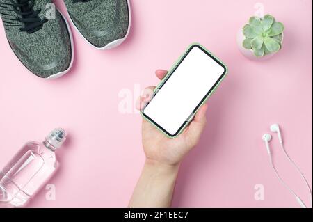 Mockup - Mann Hand hält ein Smartphone auf einem rosa Hintergrund mit Sneakers, Flasche Wasser, weißen Kopfhörern. Sportkonzept. Platzieren Sie den Text, flach legen. Stockfoto