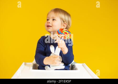 Niedlichen kleinen Jungen hält bunte Lollipop, während gegen sitzen auf Stuhl über gelben Hintergrund. Stockfoto