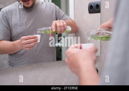 Mann gießt grüne Mundwasser aus der Flasche in die Kappe im Badezimmer. Zahnpflegekonzept. Stockfoto