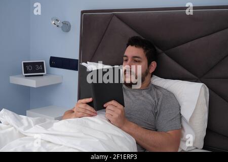 Junger latein, der am späten Morgen ein Buch im Bett las. Digitale Uhr im Hintergrund. Stockfoto