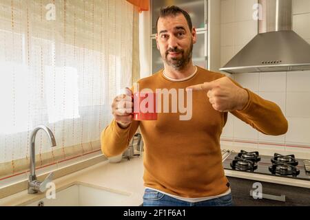 Freundlicher bärtiger Mann in einem orangefarbenen Pullover und Jeans in der Hand Eine Tasse Kaffee mit einer Hand und zeigt auf Der Becher mit der anderen Hand beim Betrachten Stockfoto
