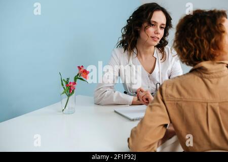 Ein hübscher Arzt, der sich in ihrem Büro um eine Patientin kümmert und sie ansieht. Auf blauem Hintergrund. Sie trägt weiße Robe und Stethoskop. Stockfoto