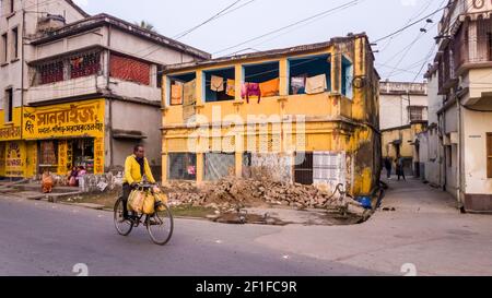 Murshidabad, Westbengalen, Indien - Januar 2018: Ein Inder in einem gelben Hemd fährt mit dem Fahrrad an den gelben Wänden von alten Häusern auf den Straßen vorbei o Stockfoto