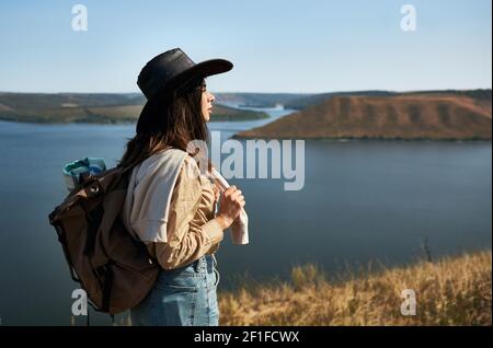 Charmante weibliche Wanderer mit Rucksack auf dem grünen Berg stehen und bewundern Sie die malerische Aussicht auf den Dniester Fluss. Aktives Hobby an der frischen Luft. Stockfoto