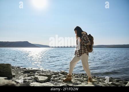 Positive Frau mit braunem Haar, die mit Rucksack am Fluss Dniester im Nationalpark Podillya Tovtry entlang läuft. Touristen verbringen Freizeit in der Natur. Aktiver Lebensstil. Stockfoto