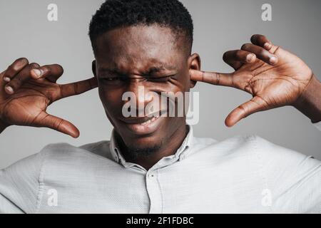 Schöner lächelnder afrikanischer Mann im weißen Hemd, der seine Ohren bedeckt, Kopfschmerzen durch lauten Lärm, isoliert auf Hintergrund Stockfoto