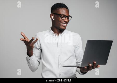 Schöner lächelnder junger afrikanischer Geschäftsmann, der Laptop auf grauem Hintergrund isoliert hält und per Videoanruf mit Kollegen spricht Stockfoto