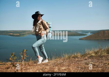Weibliche Wanderin im Cowboyhut, die mit Rucksack auf einem grünen Hügel in der Nähe des Dniester Flusses spazieren geht. Attraktive Frau mit aktiven Wochenenden an der frischen Luft in der Region Bakota. Stockfoto