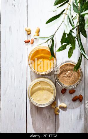 Das moderne Konzept von Wellness und Veganismus. Gläser mit Mandel-, Cashew- und Erdnussbutter auf einem weißen Holztisch mit Olivenzweig. Bio, hausgemacht Stockfoto