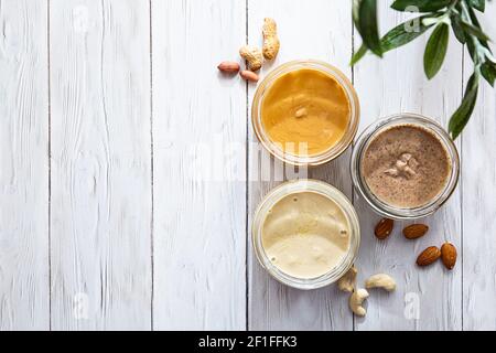 Das moderne Konzept von Wellness und Veganismus. Gläser mit Mandel-, Cashew- und Erdnussbutter auf einem weißen Holztisch mit Olivenzweig. Bio, hausgemacht Stockfoto