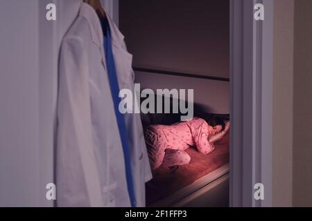 Ärztin schläft zu Hause nach der Arbeit, medizinische Uniform auf einem Kleiderbügel im Schlafzimmer Stockfoto