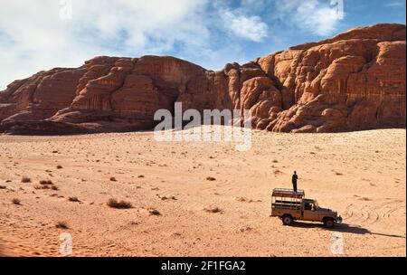 Staubige Wüste mit felsigem Massiv und blauem Himmel oben, Geländewagen im Vordergrund mit jungem Mann (unerkennbar, von hinten gesehen) auf dem steht Stockfoto