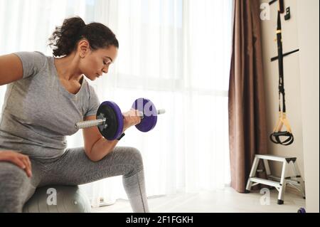 Junge afroamerikanische Frau tut Bizeps Übungen mit Hanteln an Fitnessraum auf Sportball sitzen Stockfoto