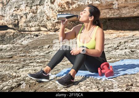 Lateinische Frau, mittleren Alters, ausruhen, Kraft zurückgewinnen, essen, Trinkwasser, nach einem Fitness-Studio-Sitzung, Kalorien zu verbrennen, fit zu halten, im Freien am Meer Stockfoto