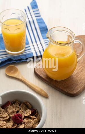 Luftaufnahme von Müslischale, Löffel, Krug und Glas mit Orangensaft, auf weißem Holz, vertikal Stockfoto