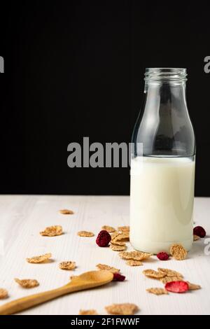 Blick auf Milchflasche auf weißem Holztisch mit Getreide und Löffel, schwarzer Hintergrund, vertikal, mit Kopierraum Stockfoto