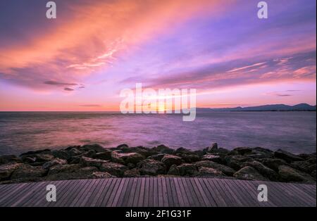 Bunte Ultra Violett dunkel Sonnenuntergang über dem Meer und mit einem Holzsteg und Felsen im Vordergrund. Stockfoto