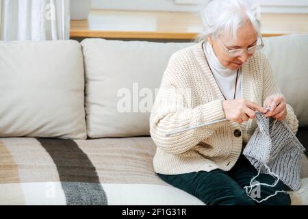Nahaufnahme einer älteren Frau, die sich mit dem Stricken auf dem Sofa zu Hause beschäftigt. Sie hat graues Haar und Hyperopie-Brille Stockfoto