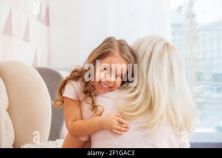 Kleine Mädchen mit kaukasischem Aussehen umarmt ihre Mutter zärtlich Ein helles Wohnzimmer im skandinavischen Stil Stockfoto