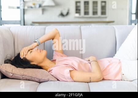 Junge asiatische Mischrassenfrau, die an Menstruationsschmerzen leidet, sich krank fühlt, Bauch hält, Kopf berührt, Bauchkrämpfe und Kopfschmerzen während der Periode erlebt, auf der Couch liegend Stockfoto