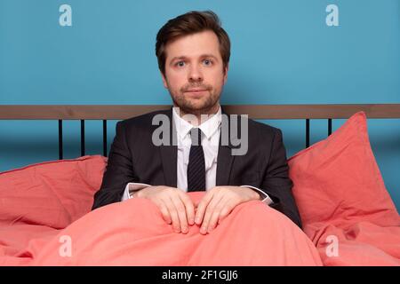 Müder Mann im Anzug sitzt am Bett und arbeitet zu Hause. Studioaufnahme Stockfoto