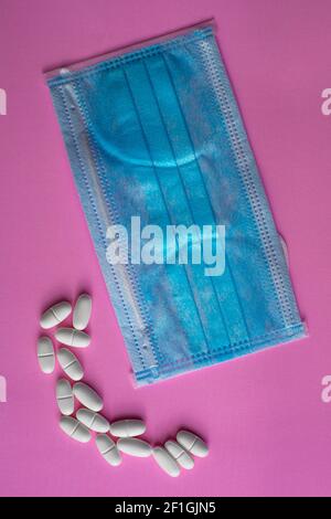 Medizinische blaue Schutzmaske und weiße Tabletten, Kapseln, Pillen oder Vitamine auf einem rosa Hintergrund Stockfoto