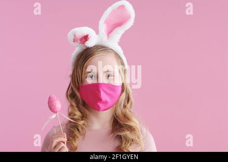 Frohe Ostern. stilvolles Kind mit langen welligen blonden Haaren mit rosa Maske, Hasenohren und osterei vor rosa Hintergrund. Stockfoto