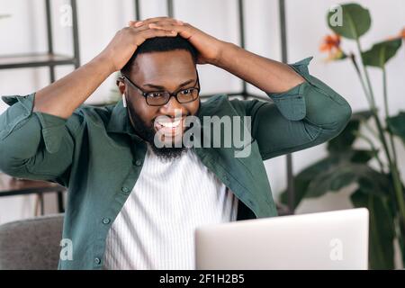 Zufrieden überrascht afroamerikanischer Kerl, sitzt an seinem Schreibtisch, hält seinen Kopf mit den Händen, schaut auf einen Laptop, erhielt eine unerwartete Nachricht oder Nachricht, sah eine Bilanz, guter Gewinn Stockfoto