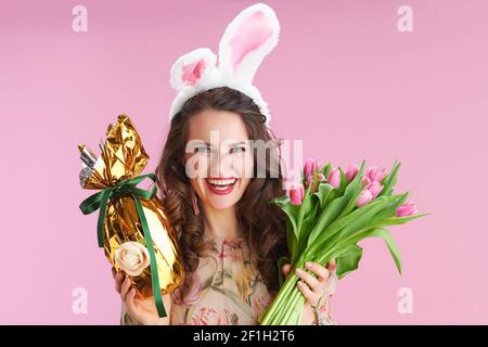 Junge Frau im Blumenkleid mit Tulpenstrauß, Hasenohren und goldenem osterei auf rosa Hintergrund. Stockfoto