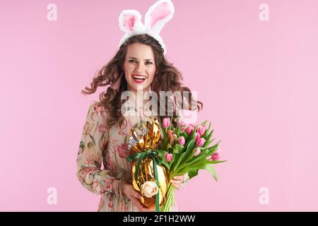 Trendige Frau mittleren Alters in Blumenkleid mit Tulpen Bouquet, Hasenohren und goldenen osterei isoliert auf rosa Hintergrund. Stockfoto