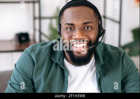 Nahaufnahme eines attraktiven, selbstbewussten bärtigen afroamerikanischen Callcenter-Betreibers oder Geschäftsführers. Schwarzer Geschäftsmann in Headset und Casual Shirt schaut auf die Kamera und freundlich lächelnd Stockfoto