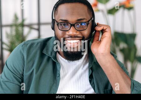 Nahaufnahme eines attraktiven, selbstbewussten bärtigen afroamerikanischen Callcenter-Betreibers oder Geschäftsführers. Schwarzer Geschäftsmann in Headset und stylish Shirt sieht auf die Kamera und freundlich Stockfoto