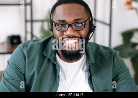 Webcam-Ansicht eines gutaussehenden selbstbewussten bärtigen afroamerikanischen Angestellten, Betreiber des Call Centers. Schwarzer Geschäftsmann in einem Headset und Brille schaut auf die Kamera und freundlich lächelnd Stockfoto