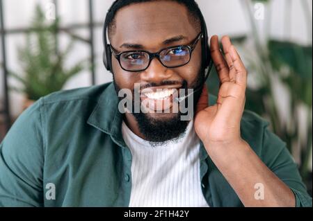 Webcam-Ansicht eines gutaussehenden selbstbewussten bärtigen afroamerikanischen Angestellten, Betreiber des Call Centers. Schwarzer Geschäftsmann in einem Headset und Brille schaut auf die Kamera und freundlich lächelnd Stockfoto