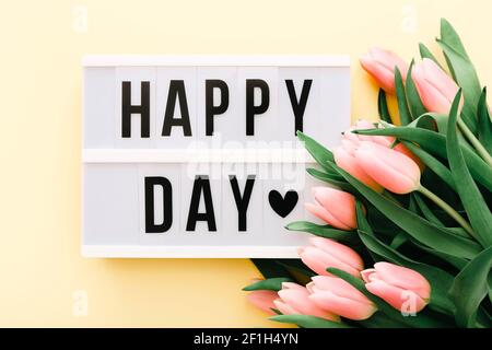 Happy Day Worte auf Leuchtkasten mit Bouquet von rosa Tulpen auf hellgelben Hintergrund. Stockfoto